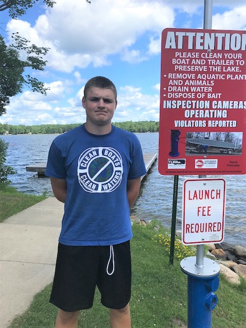 Clean Boats Clean Waters volunteer standing near boat landing with informational sign indicating best practices